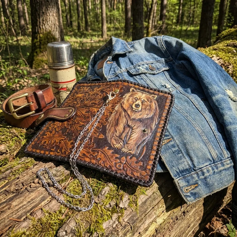 Carved grizzly bear leather biker wallet displayed on a mossy log with a denim jacket and thermos in the woods.