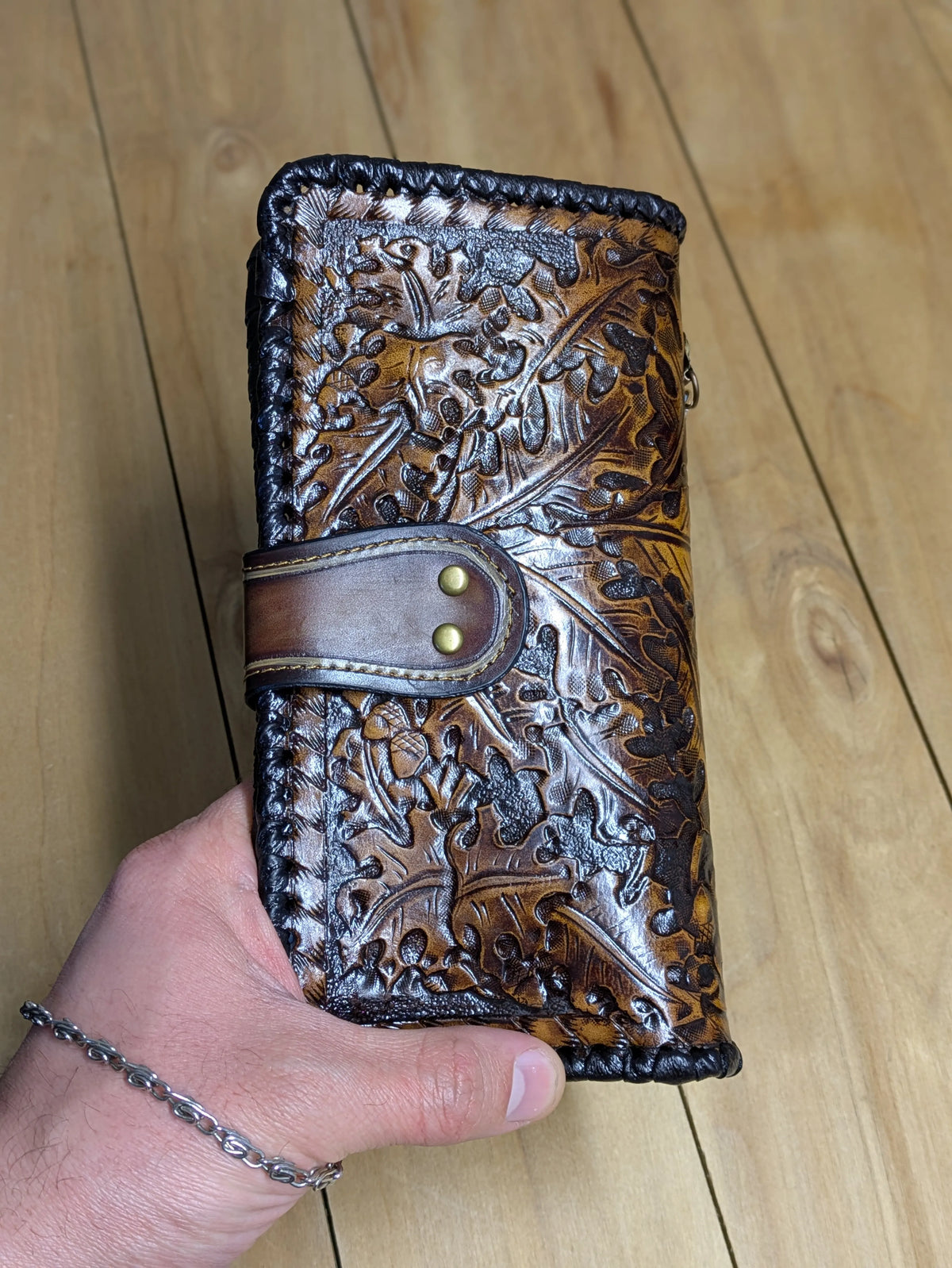 Close-up of a hand holding a folded Western leather wallet, highlighting the detailed oak leaf tooling and snap closure.