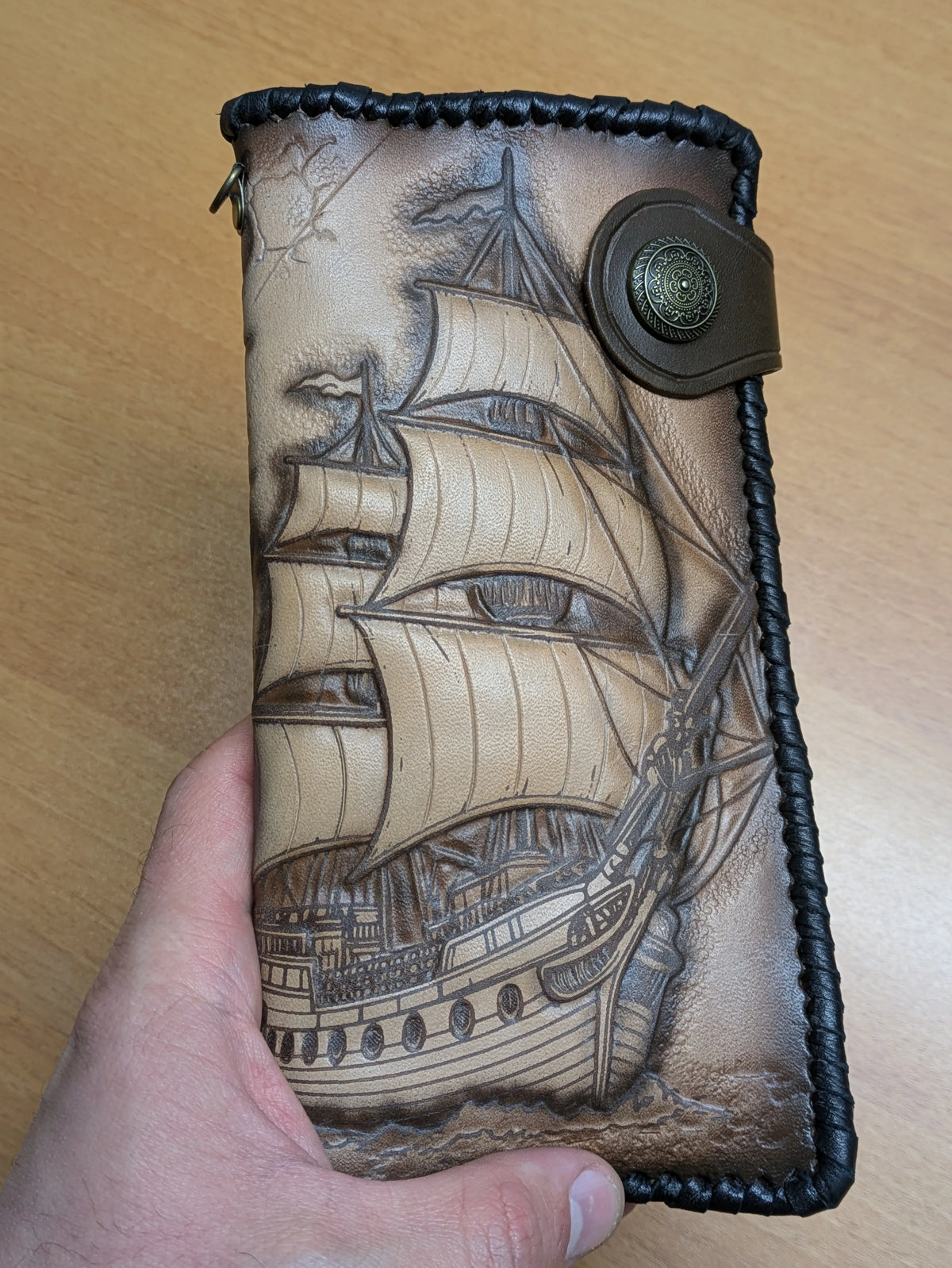 Close-up of hand-tooled leather wallet cover showing intricate details of a vintage frigate ship and decorative whip-stitched edges.