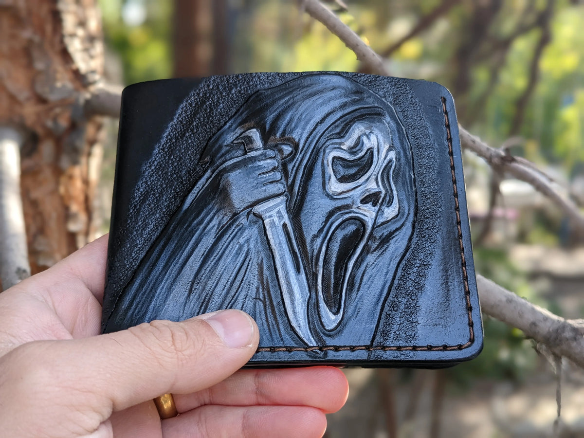 A hand holding the folded black leather Ghostface wallet outdoors, showing the textured carving against a blurred background.