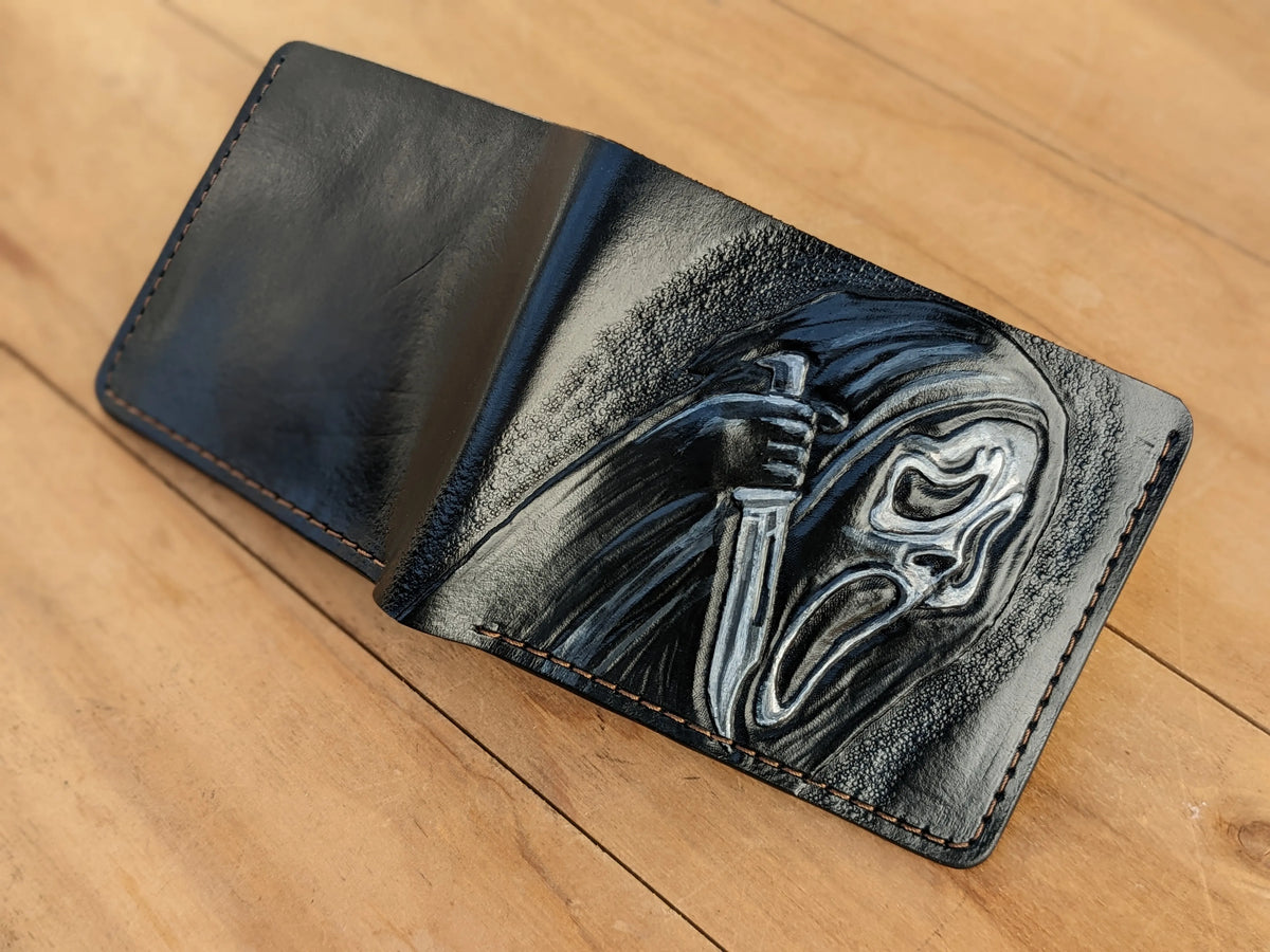 Hand-tooled leather bifold wallet laid open flat on wood, showing a smooth black back panel and a carved Ghostface front design.