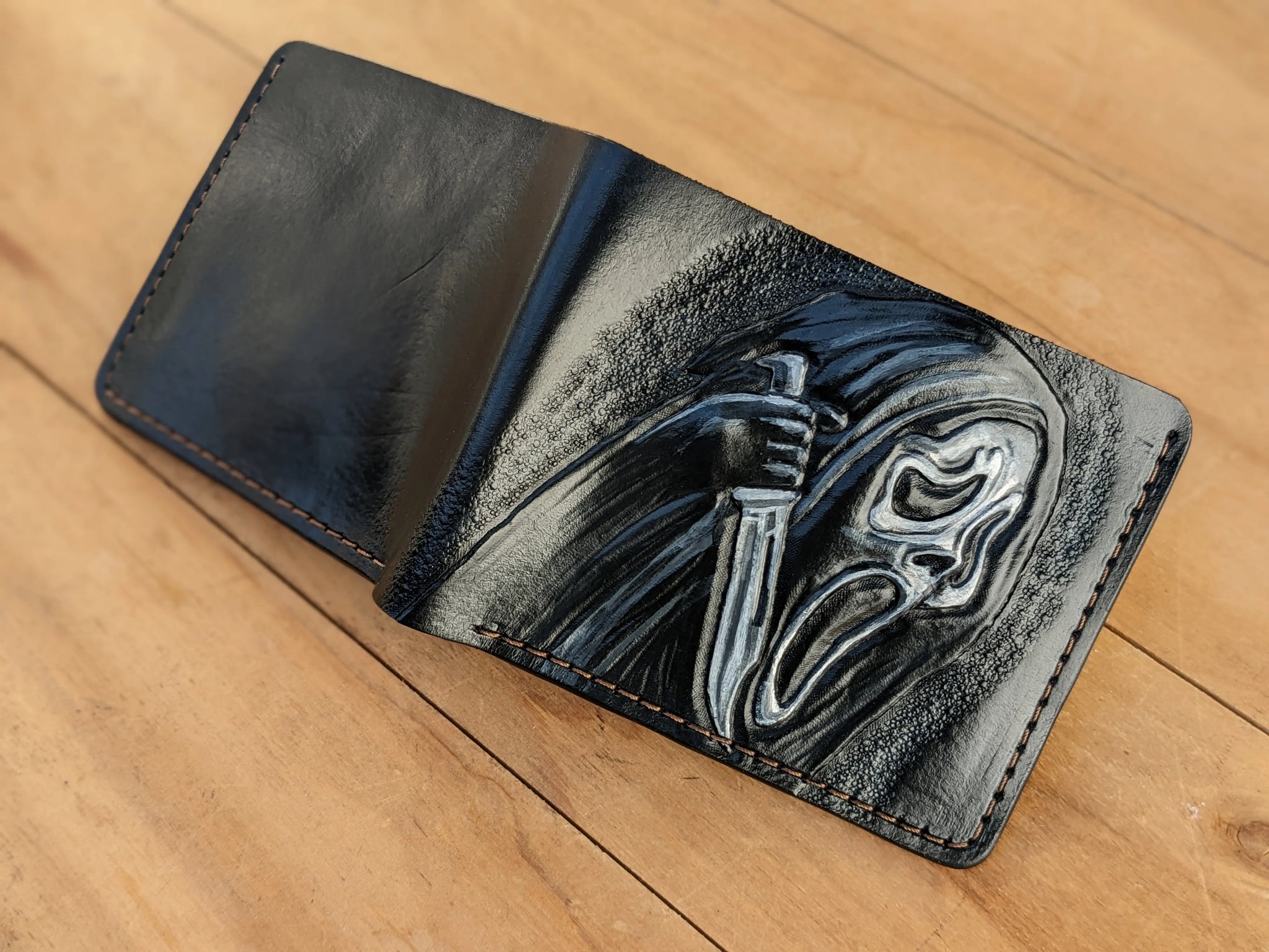 Hand-tooled leather bifold wallet laid open flat on wood, showing a smooth black back panel and a carved Ghostface front design.