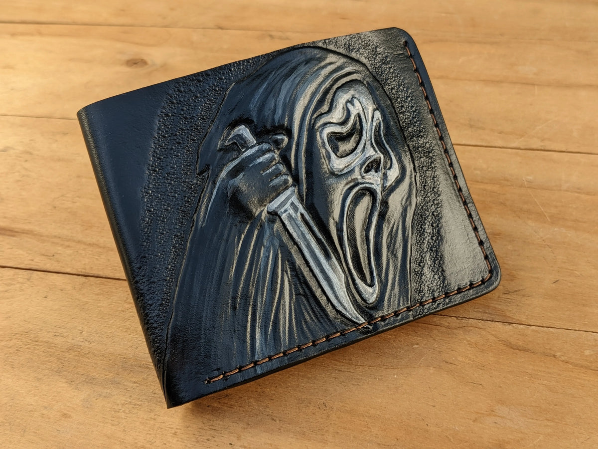 Folded black leather wallet on a wooden surface, showcasing the hand-painted highlights on the hand-tooled Ghostface carving.