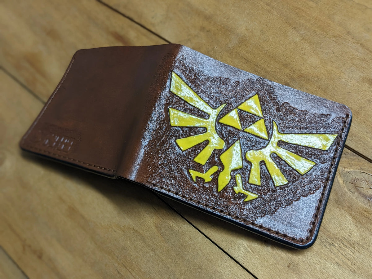 Opened Theodoros full-grain leather Zelda wallet showing hand-tooled gold Hylian Crest design on a natural wooden background.