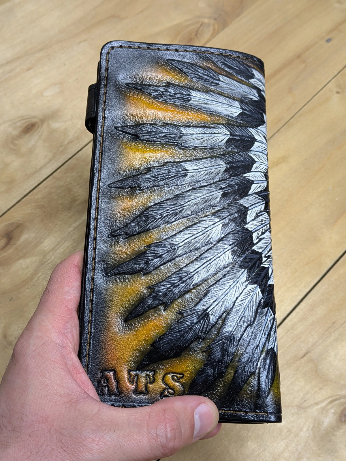 Back view of a Indian skull custom long leather wallet held by hand, showing the intricate detail of the hand-tooled feathers.