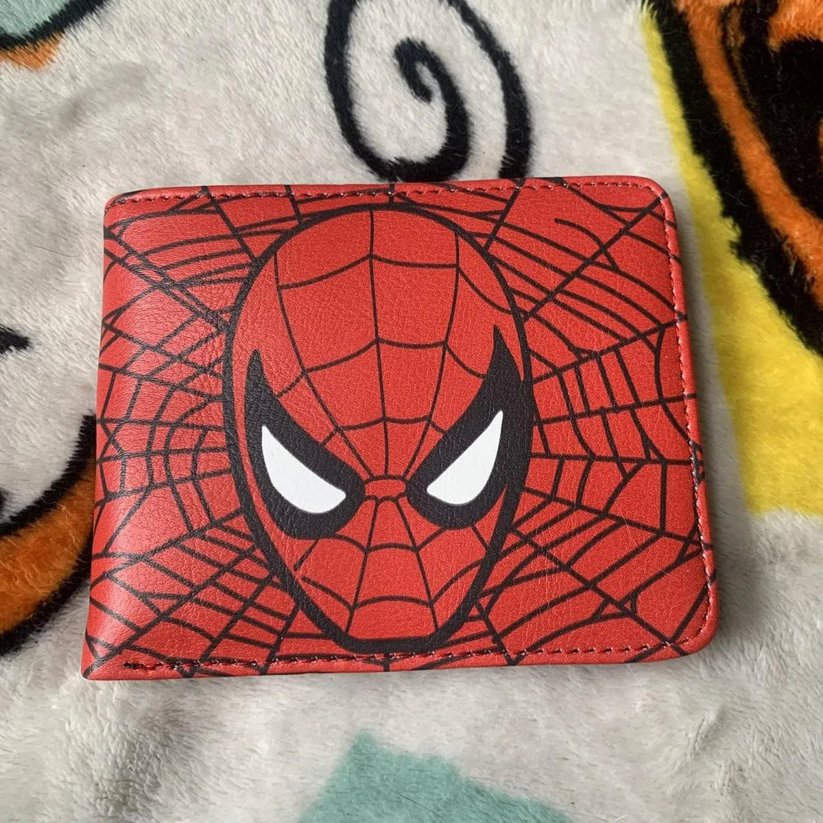 Front view of a red Marvel Spiderman bifold wallet with web pattern graphics, displayed on a soft fleece blanket.